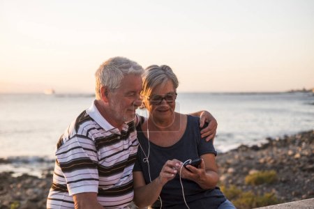 couple of senior at the beach together listen music with the same phone and the same song - woman with glasses and retired man enjoy alone - sea and rocks at the background - sunset moment 