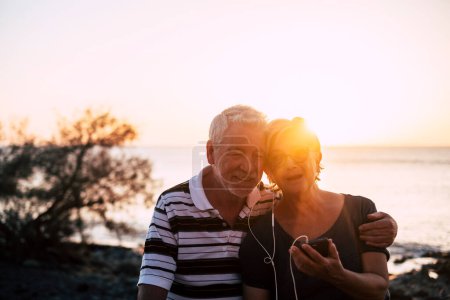 Seniorenpaar hockt bei Sonnenuntergang am Strand - Rentnerpaar hört gemeinsam Musik mit denselben Kopfhörern  