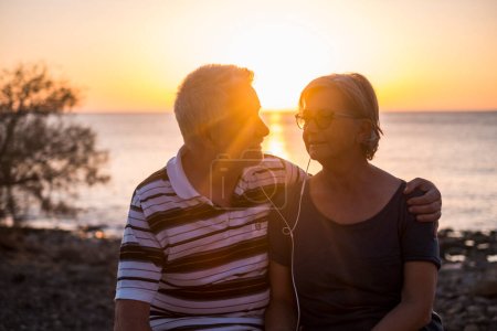 Seniorenpaar umarmt sich am Strand bei Sonnenuntergang - Rentnerpaar hört gemeinsam Musik mit demselben Kopfhörer