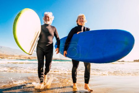 Ein paar Senioren am Strand mit schwarzen Neoprenanzügen, die ein Surfbrett in der Hand halten, um am Strand zu surfen - aktive ältere und pensionierte Menschen, die in ihrem Urlaub oder ihrer Freizeit gemeinsam fröhliche Aktivitäten ausüben 