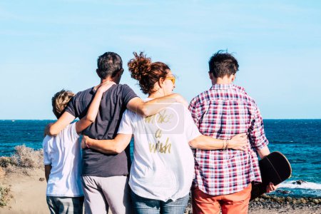 Photo for Happy family together and hugged at the beach n front and looking at the sea - four persons - child and teenager - millennial with skate board in his arm - Royalty Free Image