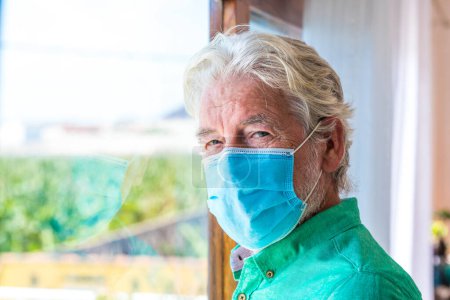 Photo for Portrait of one old and mature man looking at the camera wearing mask to prevent coronavirus indoors - lockdown lifestyle at home - Royalty Free Image