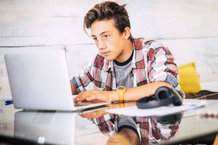 beautiful teenager focus for his studies doing homework at home on the table with laptop or computer - headphones on the table - indoor lifestyle concept - guy writing and reading