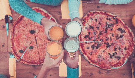 Foto de Primer plano de las manos con vasos con cerveza y zumo de naranja tintineo juntos en el centro de la mesa - dos pizzas grandes en la mesa de madera - Imagen libre de derechos