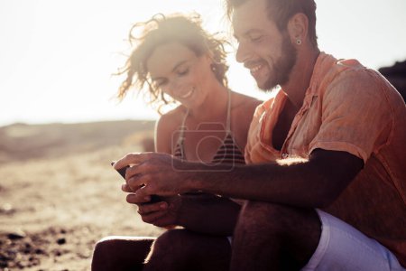 couple of adults at the beach talking and looking at the phone of the woman sitting on the rocks - woman in bikini looking at her phone and a man looking at the same phone at the sunset 