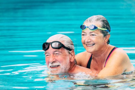 Foto de Pareja de dos ancianos abrazados en el agua de la piscina - hombre y mujer activos haciendo ejercicio juntos en la piscina - abrazados con amor - usando gafas - Imagen libre de derechos