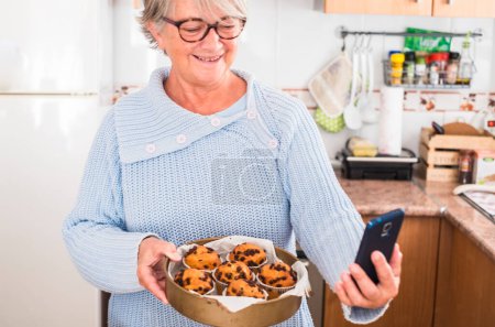 grandma holding a lot of muffins while take her a selfie in the kitchen - woman happy and enjoying - wearing glasses and blue sweater 