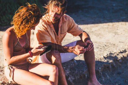 Foto de Pareja de adultos en la playa hablando y mirando el teléfono de la mujer sentada en las rocas - mujer en bikini mirando su teléfono y un hombre mirando el mismo teléfono - Imagen libre de derechos