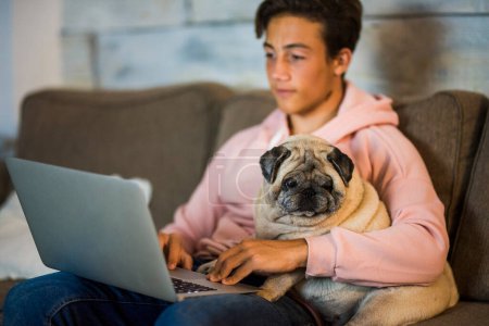 Teenager zu Hause mit Laptop auf dem Sofa, neben ihm ein Mops - beste Freunde auf dem Sofa umarmt 