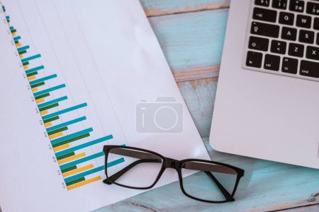 close up of laptop or computer pc with glasses and graphs and statistics on a blue wood table - nobody at the photo 