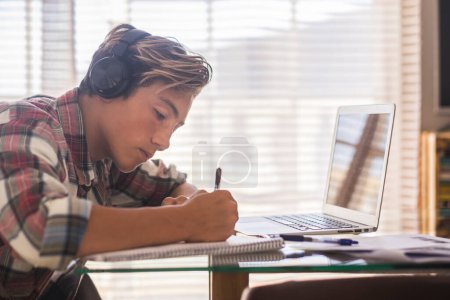 Caucasian teenager indoor doing homework on the table at home - blonde guy writing and reading in his laptop or computer to get great scores