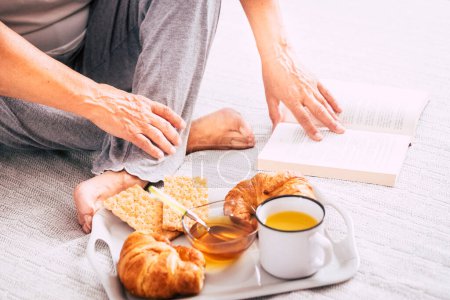 Ältere Frau sitzt auf dem Bett und liest ein Buch und isst Croissant und Cracker und trinkt Orangensaft - Rentnerin frühstückt allein und isoliert am Morgen  
