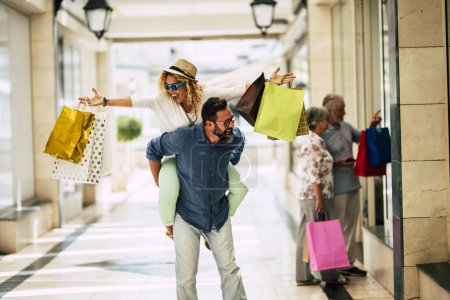 glückliche Familie geht gemeinsam einkaufen - zwei Erwachsene haben Spaß und zwei Senioren im Hintergrund schauen sich die Geschäfte an - Mann hält eine Frau auf dem Rücken 