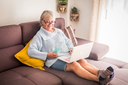 Technik, Alter und Menschen-Konzept - glückliche Seniorin in Brille mit Laptop und Glas vom Zuhause  