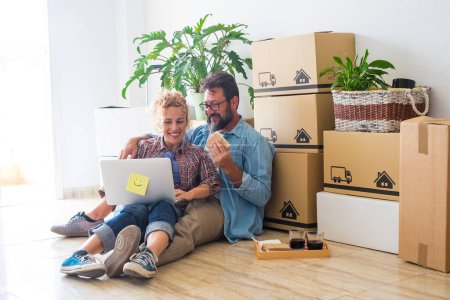 Foto de Pareja de dos adultos felices y personas sentadas en el suelo usando su computadora portátil juntos y comiendo un bocadillo después de hacer una hipoteca - Imagen libre de derechos
