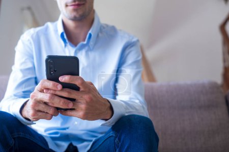 Photo for Hands of man sitting on sofa and text messaging using mobile phone at home. Caucasian guy browsing social media content or chatting online on smartphone while relaxing on couch - Royalty Free Image