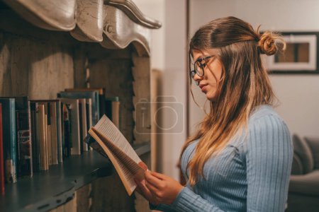 Un mignon et attrayant portant des lunettes de lecture et à la recherche d'un livre intéressant à la maison à l'intérieur. Jeune femme se détendre seul la nuit lecture de livres 
