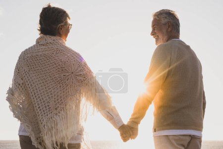 couple of two happy and active seniors having fun and enjoying together summer at the beach walking holding their hands with the sunset at the background 