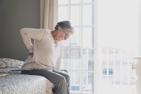 Foto de Mujer mayor cansada que sufre de dolor de espalda después del sueño, frotando los músculos rígidos, Mujer vieja sentada en la cama tocando la parte inferior de la espalda sintiendo malestar debido a la cama incómoda en casa - Imagen libre de derechos