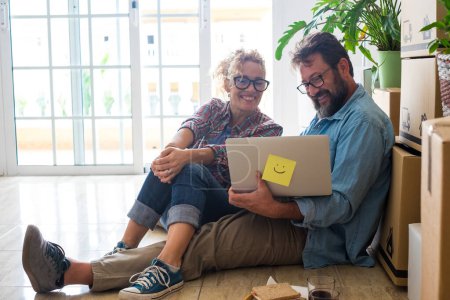Téléchargez les photos : Couple de deux adultes heureux et les gens assis sur le sol en utilisant leur ordinateur portable ensemble et de manger une collation après avoir fait une hypothèque - en image libre de droit