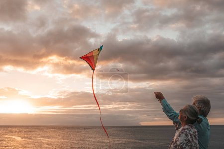 Photo for Close up and portrait of two old and mature people playing and enjoying with a flaying kite at the beach with the sea at the background with sunset - active seniors having fun - Royalty Free Image