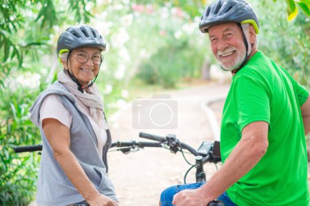 Two happy old mature people enjoying and riding bikes together to be fit and healthy outdoors. Active seniors having fun training in nature. Portrait of two senior looking at the back looking at the camera