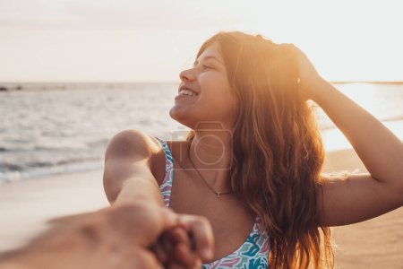 Photo for Portrait and close up of young woman holding hand of boyfriend walking on the beach smiling and having fun together. Man following girlfriend to the water of the sea enjoying. - Royalty Free Image