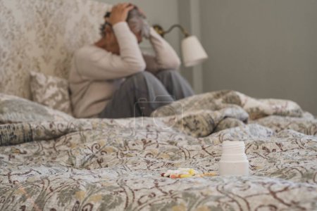 Tabletten oder Pillen auf dem Bett, im Hintergrund sitzt eine an Depressionen leidende Seniorin. Alte Dame hält ihren Kopf in starken Migräne-Schmerzen, fühlt sich unruhig und unwohl im Schlafzimmer mit Medikamenten im Vordergrund  