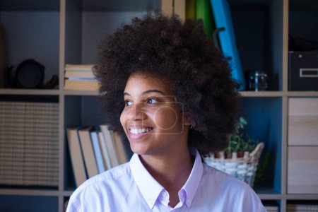Attractive dark skinned young woman with curly Afro hairstyle looking out through window. Thoughtful african american businesswoman smiling and looking away at office. Female lady dreaming about future 