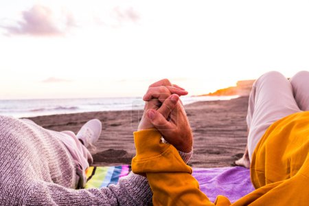 Foto de Primer plano de dos manos unidas en la playa con la puesta de sol al fondo disfrutando del verano y divirtiéndose juntas. Un par de personas en la arena - Imagen libre de derechos