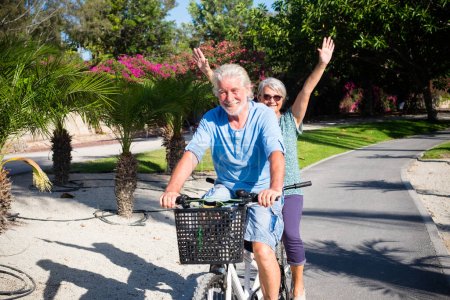 Téléchargez les photos : Beau et mignon couple de mature et vieille femme et homme chevauchant ensemble un vélo double dans un parc verdoyant avec des fleurs roses à l'arrière-plan. Aîné actif s'amuser avec tandem - en image libre de droit