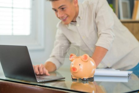 Man sit at desk manage expenses, calculate expenditures, pay bills online use laptop, makes household finances analysis, close up focus on pink piggy bank. Save money for future, be provident concept 