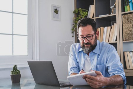 Photo for One young man working at home in the office with laptop and notebook taking notes talking in a video conference. One businessman calling communicating - Royalty Free Image