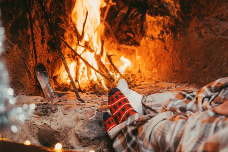 Low section legs of husband and wife covered in socks and warm blanket relaxing in front of burning fireplace during winter christmas holiday. Pair of feet in woolen socks by the Christmas fireplace 