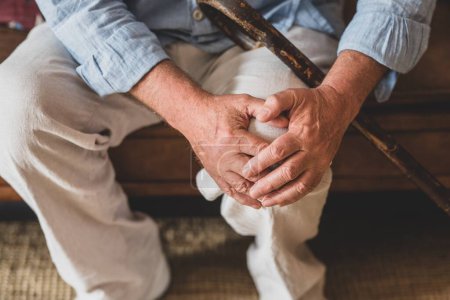 Großaufnahme eines älteren Mannes mit Knieproblemen, der zu Hause auf dem Sofa sitzt und die Knie hält. Älterer Mann mit starken Knieschmerzen sitzt mit Gehstock im Wohnzimmer. 