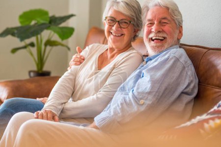 Portrait of cheerful senior couple embracing while sitting on sofa and smiling. Elderly happy couple relaxing and posing in front of camera sitting in living room.