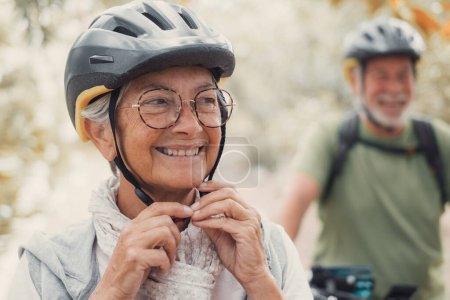 Porträt einer alten Frau, die lächelt und die Natur genießt, während ihr Mann lacht. Kopfschuss einer reifen Frau mit Brille, die sich gesund fühlt. Senior setzt Helm auf, um mit dem Fahrrad unterwegs zu sein.  