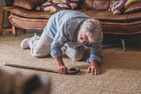 Älterer Senior liegt nach Sturz mit Gehstock neben Couch auf Teppich im heimischen Wohnzimmer am Boden. Alter Mann leidet unter Schmerzen und kämpft nach Sturz im Haus ums Aufstehen