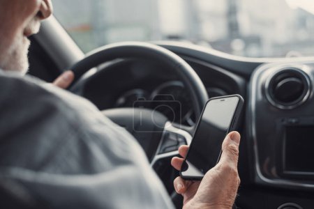 Photo for Man looking at mobile phone while driving a car. Old pensioner person distracting texting and chatting. - Royalty Free Image