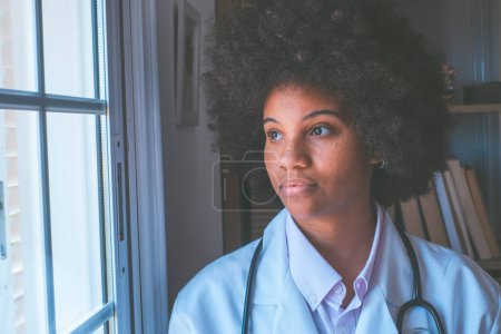 Photo for Thoughtful female african american doctor with stethoscope looking outside her clinic window. Serious contemplative frontline heathcare worker with curly hair admiring view from window - Royalty Free Image
