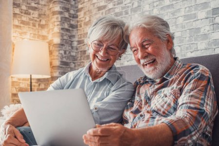 Linda pareja de ancianos sentados en el sofá utilizando el ordenador portátil juntos de compras y navegar por la red. Dos personas maduras en el salón disfrutando de la tecnología. Retrato de seniors riendo en el amor. 