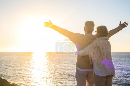 Téléchargez les photos : Couple de deux heureux aînés ad personnes âgées matures à la plage en regardant la mer et le soleil avec les bras ouverts sentiment de liberté. Concept de liberté et mode de vie - en image libre de droit