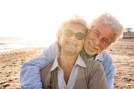 Téléchargez les photos : Portrait de couple de personnes âgées et matures profitant de l'été à la plage regardant vers la caméra souriant et s'amusant avec le coucher de soleil en arrière-plan. Deux aînés actifs voyageant à l'extérieur. - en image libre de droit