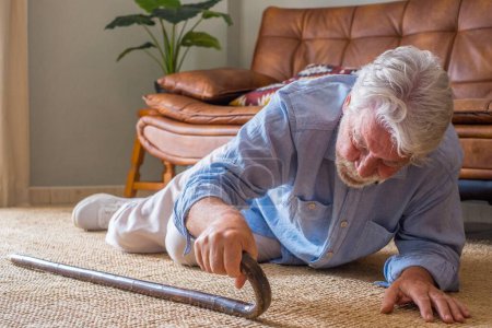 Älterer Senior liegt nach Sturz mit Gehstock neben Couch auf Teppich im heimischen Wohnzimmer am Boden. Alter Mann leidet unter Schmerzen und kämpft nach Sturz zu Hause ums Aufstehen