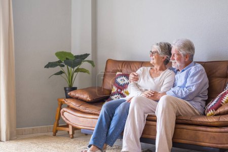 Photo for Senior couple relaxing on sofa at home and looking out through window on a bright sunny day. Happy old husband and wife looking away and admiring view sitting on couch at modern house - Royalty Free Image