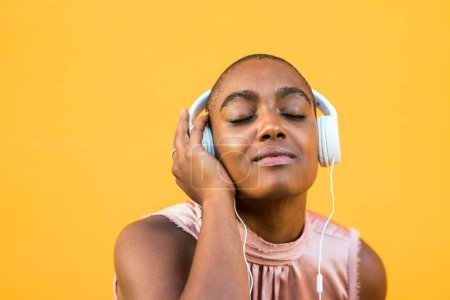 Photo for Dark skinned woman listening to music on headphones with eyes closed over yellow background. African american lady enjoying her favourite song using wired audio equipment. Technology and entertainment - Royalty Free Image