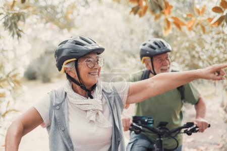 Photo for Two happy old mature people enjoying and riding bikes together to be fit and healthy outdoors. Active seniors having fun training in nature. Portrait of one old man smiling in a bike trip with his wife. Woman indicating something and looking at it. - Royalty Free Image