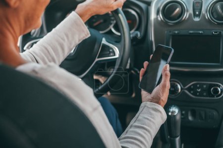 Photo for Portrait of one old woman using phone in car while driving in the road risk causing an accident. Close up of hands holding smartphone surfing online texting and chatting. - Royalty Free Image