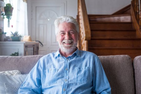 Téléchargez les photos : Portrait de l'homme âgé heureux assis sur le canapé dans le salon de la maison en face de l'escalier en bois. Vieux mâle souriant tout en se relaxant sur le canapé et en regardant la caméra - en image libre de droit