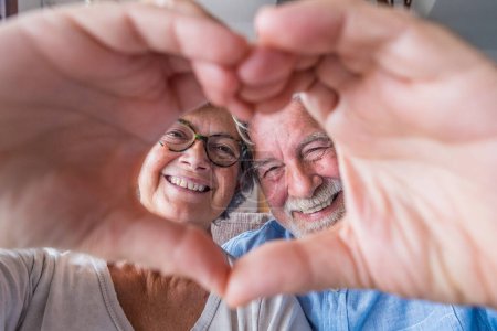 Close up portrait happy sincere middle aged elderly retired family couple making heart gesture with fingers, showing love or demonstrating sincere feelings together indoors, looking at camera. 